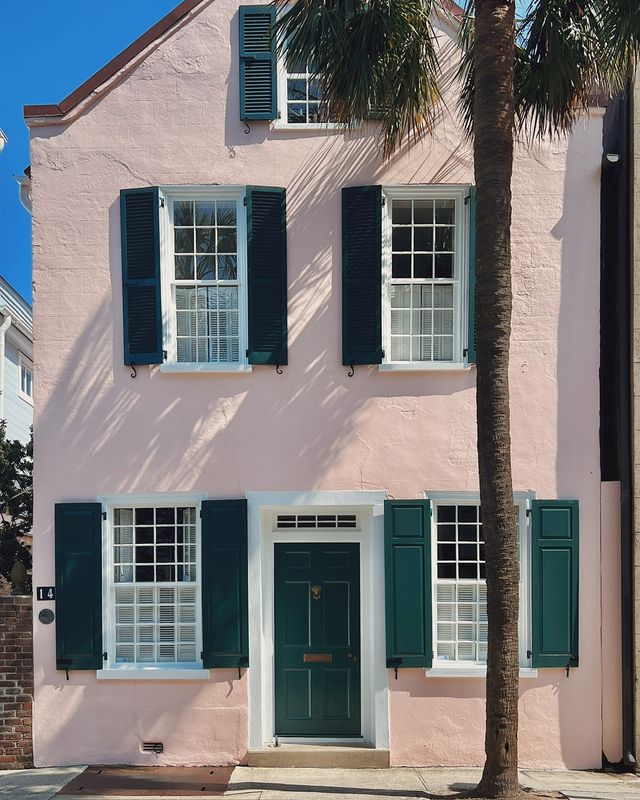 Charming pastel pink two-story house with dark green shutters and door, a tall palm tree casting sunlit shadows against a clear blue sky