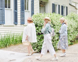 Three women in sun hats and casual summer outfits strolling past a white cottage with blue shutters and a dense white‑flowering hedge on a brick sidewalk — picturesque spring neighborhood scene.