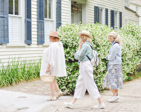 Three women in sun hats and casual summer outfits strolling past a white cottage with blue shutters and a dense white‑flowering hedge on a brick sidewalk — picturesque spring neighborhood scene.