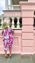 Person in a floppy green sun hat and white dress with bright pink floral print standing on stone pavement, peering over a pastel-pink balustrade of a Victorian-style building with white columns and balcony.