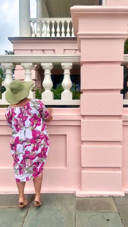 Person in a floppy green sun hat and white dress with bright pink floral print standing on stone pavement, peering over a pastel-pink balustrade of a Victorian-style building with white columns and balcony.