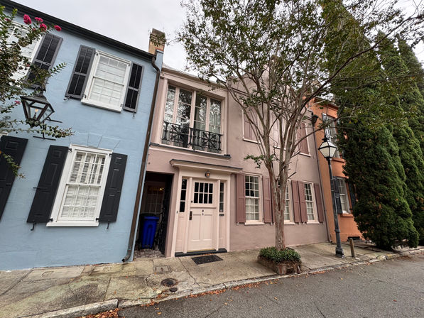 Charming pastel row houses on a narrow historic street – blue and blush facades with shuttered windows, wrought-iron balcony, sidewalk tree and classic street lamp.