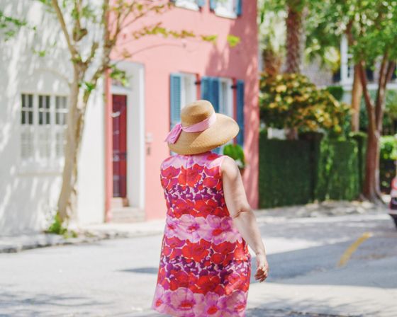 Woman in a pink-and-red floral sundress and straw sun hat walking away on a sunny residential street lined with pastel houses, blue shutters, trees, and green hedges.
