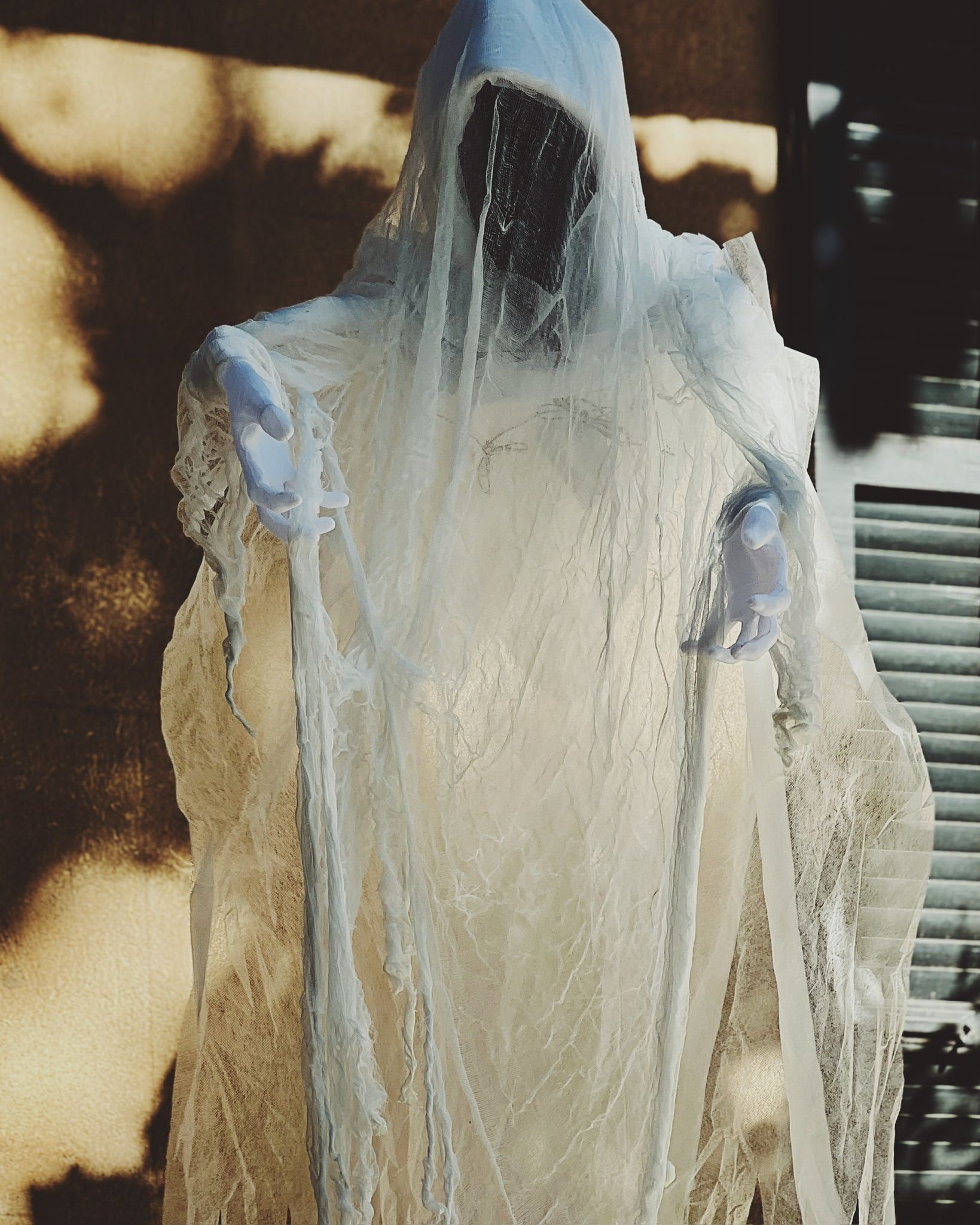 Lifesize Halloween ghost decoration draped in tattered white gauze with a dark shrouded face and outstretched gloved hands, sunlit against a house wall and shutters.