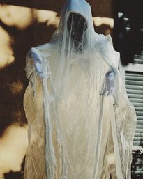 Lifesize Halloween ghost decoration draped in tattered white gauze with a dark shrouded face and outstretched gloved hands, sunlit against a house wall and shutters.