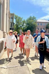 Group of friends enjoying a sunny historic downtown walking tour, dressed in summer dresses, hats and sneakers, sipping iced drinks with pastel buildings and live oak trees lining the sidewalk.