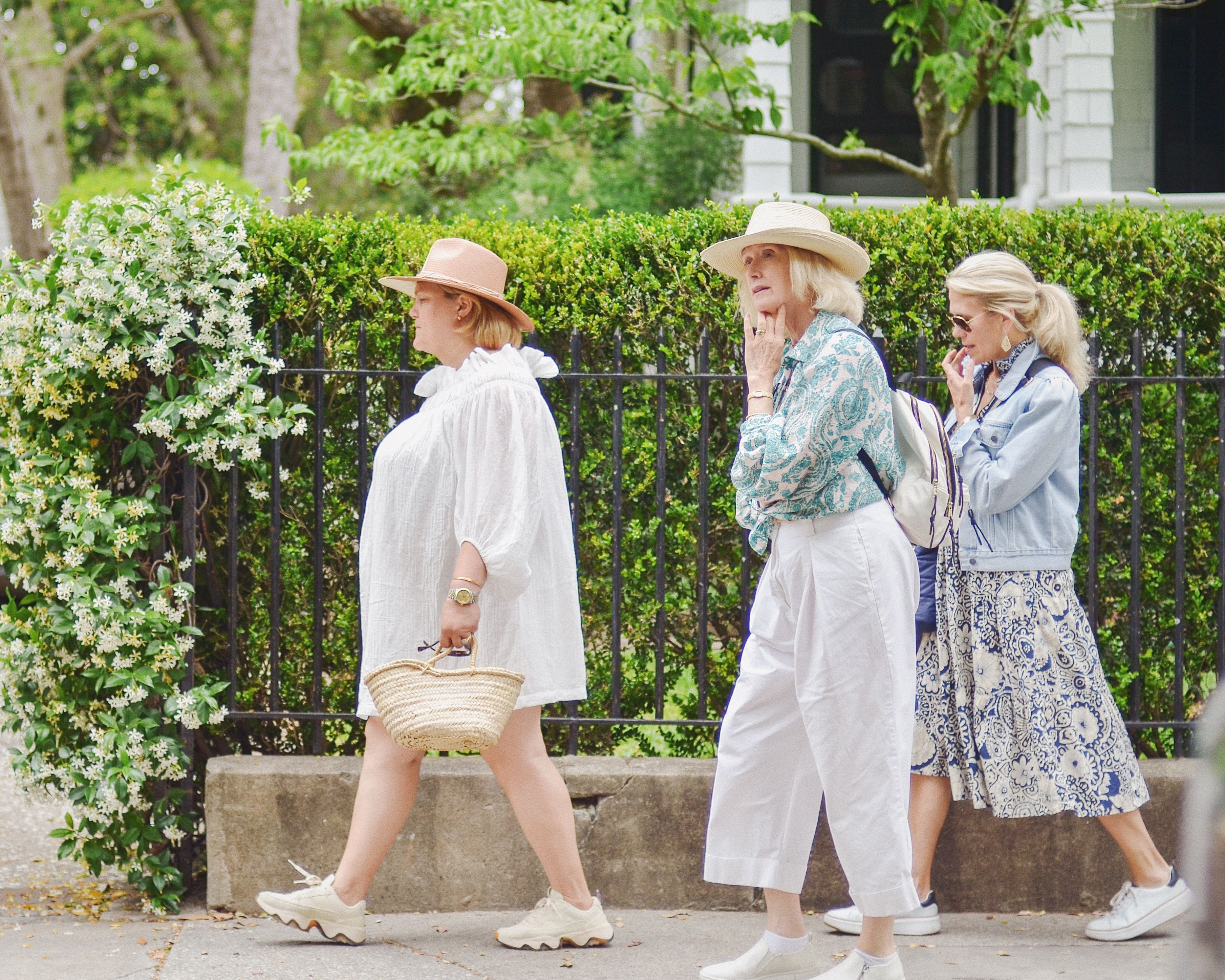 Three women strolling along a leafy residential sidewalk beside a trimmed hedge and iron fence, wearing summer outfits—straw hats, a woven basket, patterned midi skirt with a denim jacket, white wide-leg trousers and sneakers.