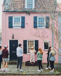 Guide in a floral dress and sun hat leading a small walking tour outside a pastel pink historic house with black shutters, tiled roof, and bare tree
