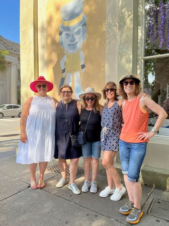 Five women smiling on a sunny historic downtown sidewalk in front of a playful wall mural, wearing summer dresses, hats and sunglasses.
