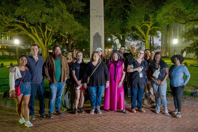 Group photo of adults at night in a historic park square with live oaks and a stone monument; one person in a bright pink cloak and witch hat stands with casually dressed tour participants during an evening walking tour.