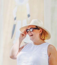 Smiling woman in a white sleeveless top and straw sun hat adjusting dark sunglasses in a sunlit outdoor summer scene