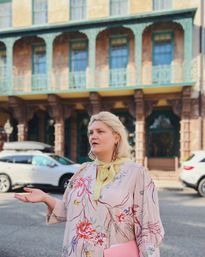 Person in a floral dress and yellow neck scarf gesturing with one hand and holding a pink folder on a sunny historic downtown street, ornate balconyed brick building and parked cars in the background
