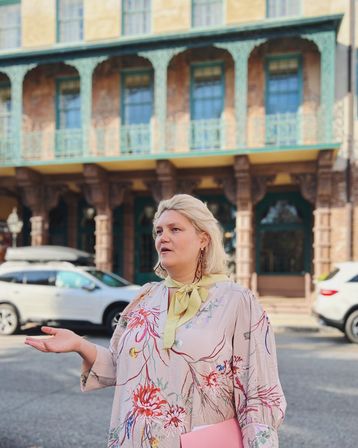 Person in a floral dress and yellow neck scarf gesturing with one hand and holding a pink folder on a sunny historic downtown street, ornate balconyed brick building and parked cars in the background