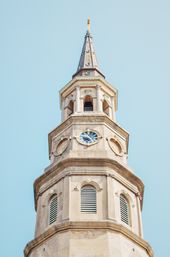 Historic pale-stone church steeple and clock tower with ornate columns, louvered windows and a pointed spire topped by a gold finial against a clear blue sky.