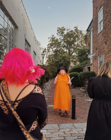 Person in a bright orange dress and black hat gesturing with papers in a narrow historic brick alley, two listeners in foreground (one with neon pink hair), cobblestone path, lamppost and trees at dusk.