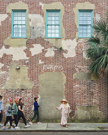 Weathered red brick building with peeling plaster and three turquoise-framed windows, a palm tree at right, and a group of pedestrians on the sidewalk passing a woman in a floral dress and wide-brim sun hat.