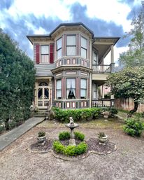 Charming two-story Victorian house with octagonal bay windows, red shutters, decorative trim and porch, front yard garden with boxwood edging and a stone birdbath under a cloudy sky