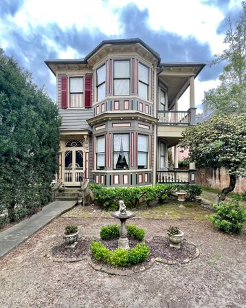 Charming two-story Victorian house with octagonal bay windows, red shutters, decorative trim and porch, front yard garden with boxwood edging and a stone birdbath under a cloudy sky