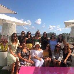 Twelve women smiling on a sunny rooftop patio under white umbrellas, wearing summer outfits and sunglasses with blue sky, puffy clouds and a pink table in the foreground.