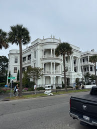 Historic white antebellum mansion with ornate columns, wraparound balconies and palm trees along East Battery, pedestrians on the sidewalk under an overcast sky.