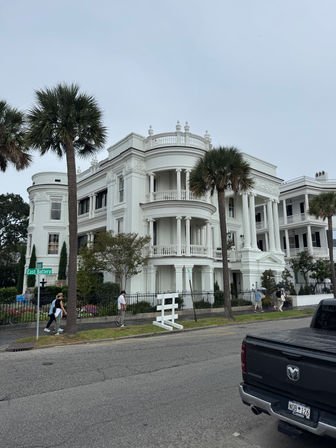 Historic white antebellum mansion with ornate columns, wraparound balconies and palm trees along East Battery, pedestrians on the sidewalk under an overcast sky.