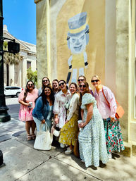 Group of smiling women in colorful spring dresses posing on a sunny historic downtown sidewalk beside a beige wall mural of a whimsical top-hatted character, with palm trees and classical columns visible in the background.