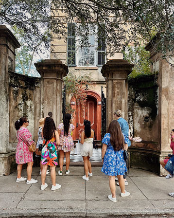Tour group admiring a historic townhouse entrance with an arched coral door, mossy stone gate, wrought-iron details and a checkerboard stoop under oak branches