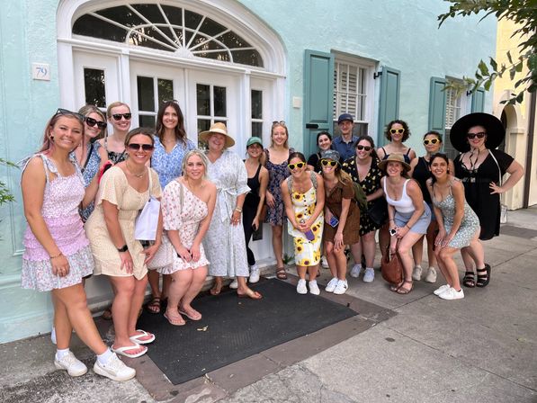 Large group of women in summer dresses and sunglasses posing and smiling outside a pastel mint-green historic building with an arched white door and teal shutters on a sunny street
