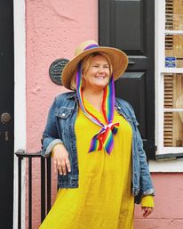 Smiling woman in a straw hat with rainbow ribbons, yellow dress and denim jacket leaning against a pink stucco wall by a black door and shuttered window.