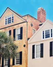 Sunlit pastel peach and blush row houses with gabled roofs, black shutters and a tall decorative chimney, palm tree in front and bright blue sky for a coastal historic charm