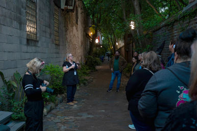 Evening guided walking tour in a narrow, lantern-lit historic brick alley — a guide speaks to a small group under a tree canopy while a photographer adjusts a camera at the side.