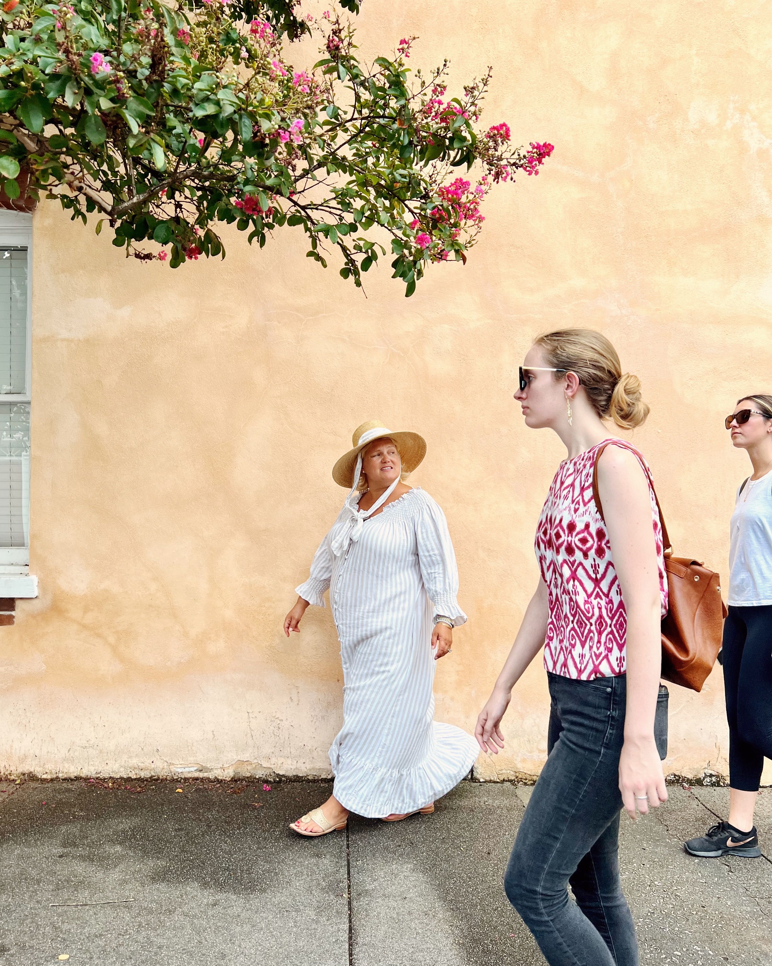 Three women strolling on a sunny sidewalk by a peach stucco wall beneath pink flowering branches — one in a striped sun dress and wide straw hat, another in a patterned sleeveless top and sunglasses, and a third in casual activewear.