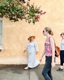 Three women strolling on a sunny sidewalk by a peach stucco wall beneath pink flowering branches — one in a striped sun dress and wide straw hat, another in a patterned sleeveless top and sunglasses, and a third in casual activewear.
