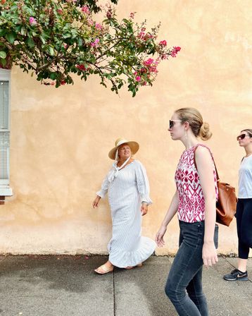 Three women strolling on a sunny sidewalk by a peach stucco wall beneath pink flowering branches — one in a striped sun dress and wide straw hat, another in a patterned sleeveless top and sunglasses, and a third in casual activewear.