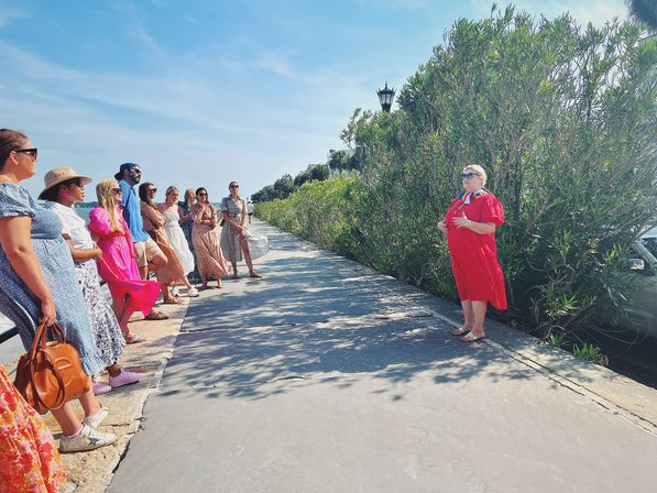 Tour group on a sunny seaside promenade listening to a guide in a bright red dress beside dense green shrubs and lampposts