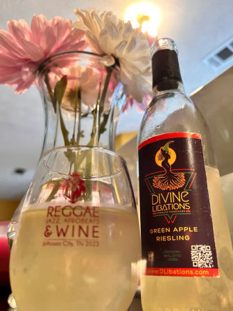 Close-up of a chilled green apple Riesling bottle and a filled wine glass with an event logo referencing Johnson City, TN, beside a vase of pink and white daisies for a bright, casual wine-tasting scene.