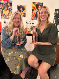 Two smiling women seated in a tasting room holding matching bottled drinks, with a counter of liquor bottles and colorful pop-art wine posters on the wall behind them.