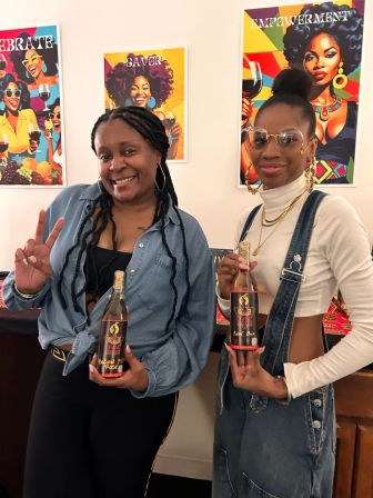 Two smiling women pose indoors holding rosé wine bottles, one flashing a peace sign, with colorful empowerment and celebratory Afrocentric posters decorating the wall behind them.