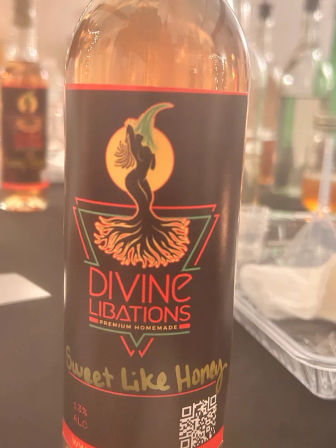 Amber liqueur bottle on a tasting table, black label with stylized mermaid silhouette against an orange moon, handwritten 'Sweet Like Honey' note and QR code.