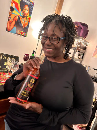 Smiling person with glasses holding a bottled beverage in a cozy home kitchen/bar area with colorful pop-art poster, wine glasses, and string lights.