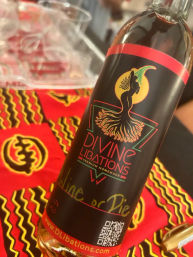 Bottle of artisanal homemade liqueur with a black label featuring a dancing silhouette and sun motif, placed on vibrant red-and-yellow patterned fabric.