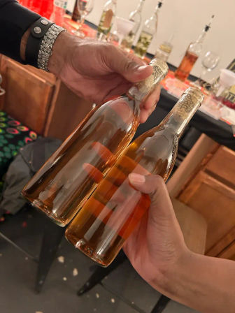 Close-up of two hands clinking amber liquor bottles with gold foil tops over a kitchen counter at a casual indoor tasting or party.