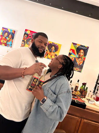 Two smiling people holding bottled beverages in an indoor tasting room, standing by a wooden bar with bottles and snacks and colorful pop-art portraits of women on the white wall behind them.