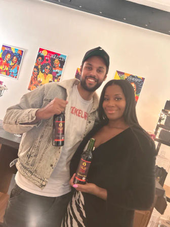 Smiling man and woman holding bottles of red wine in a bright indoor wine-tasting room with colorful pop-art posters on the wall.