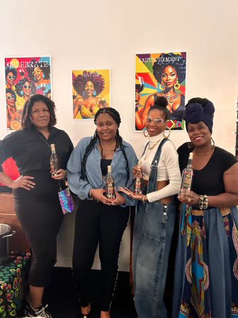 Four women posing indoors at an art-filled tasting, each holding a bottled beverage in front of colorful pop-art posters reading Celebrate, Savor, and Empowerment.
