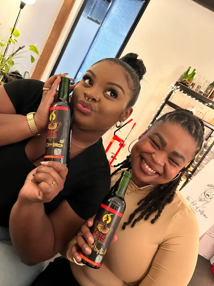 Two women smiling in a cozy indoor tasting space, each holding a dark-labeled bottle—one playfully presses it to her cheek