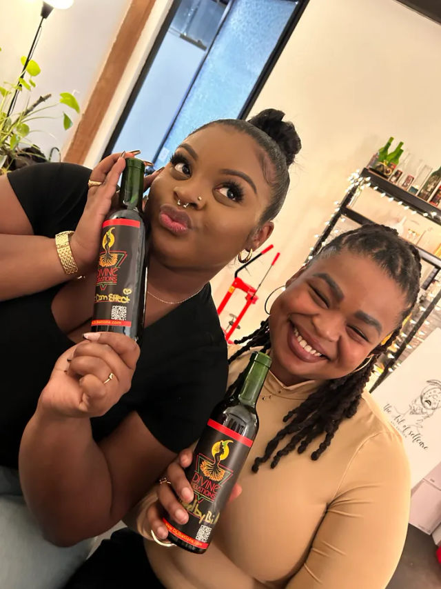 Two women smiling in a cozy indoor tasting space, each holding a dark-labeled bottle—one playfully presses it to her cheek