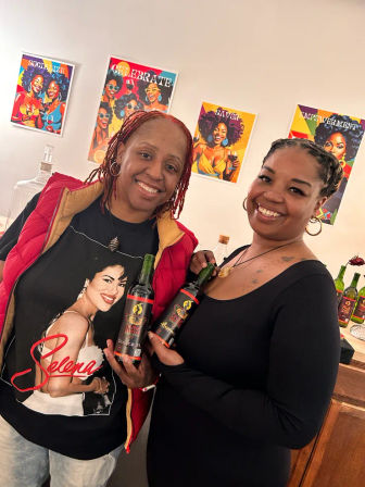 Two smiling women in a cozy tasting-room setting holding black-labeled wine bottles, standing in front of vibrant pop-art posters on the wall.