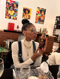 Woman at an indoor cocktail tasting playfully kissing a bottle of liqueur, seated at a table with glasses, jars and a funnel, colorful retro Afrocentric posters on the wall behind her.