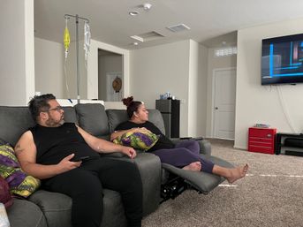 Couple relaxing on a gray sectional couch in a bright home living room, watching wall-mounted TV while one person receives at-home IV infusion from a pole with yellow and clear bags.